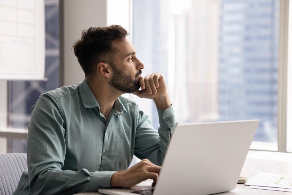 Pensive middle-aged man distracted from workflow looking out window to big city with contented smile, daydreaming about a new career